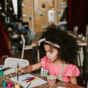 A young girl focuses on her painting in a lively art workshop setting, expressing creativity.