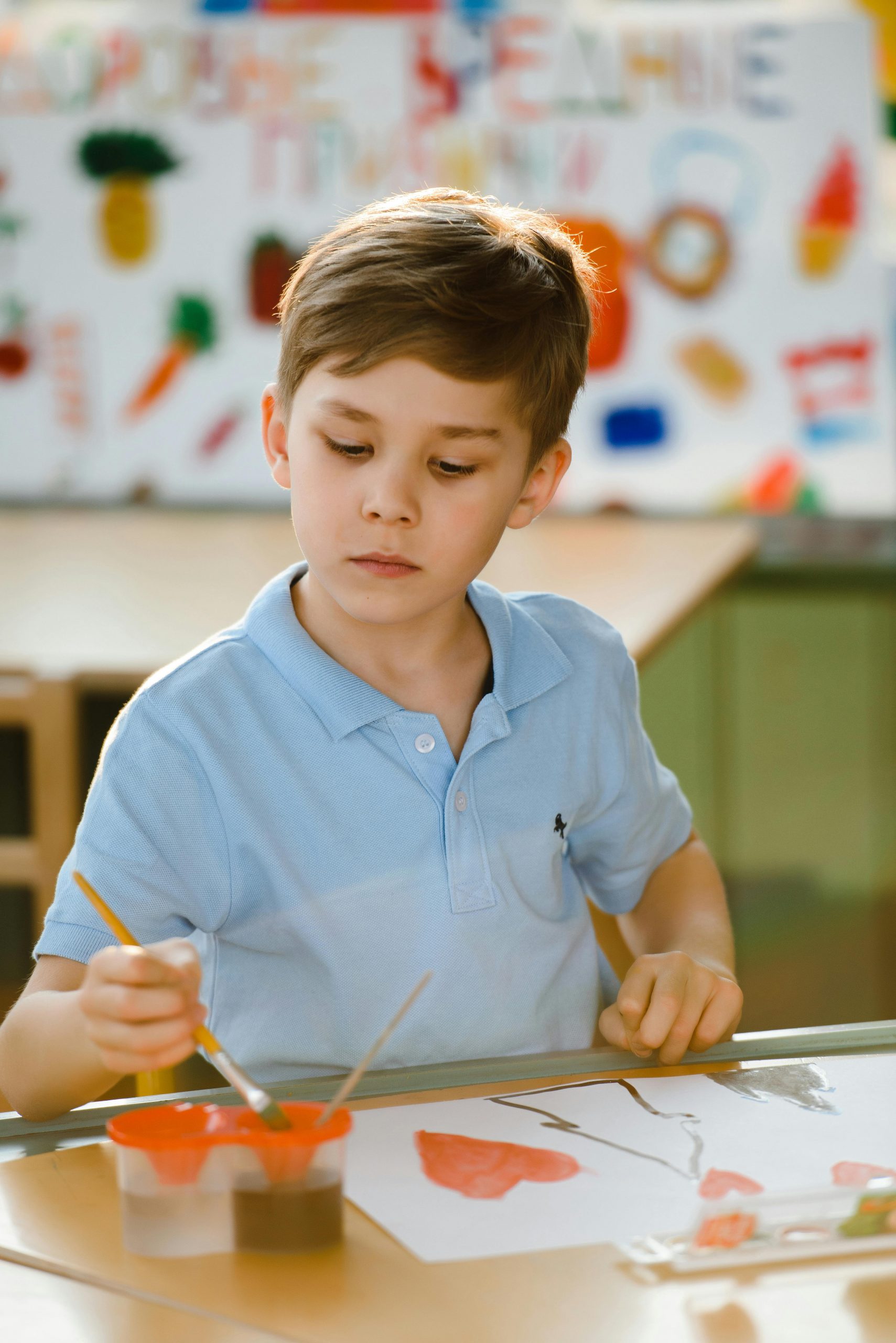 A young boy concentrating deeply on painting a creative piece of art using bright colors in a classroom.