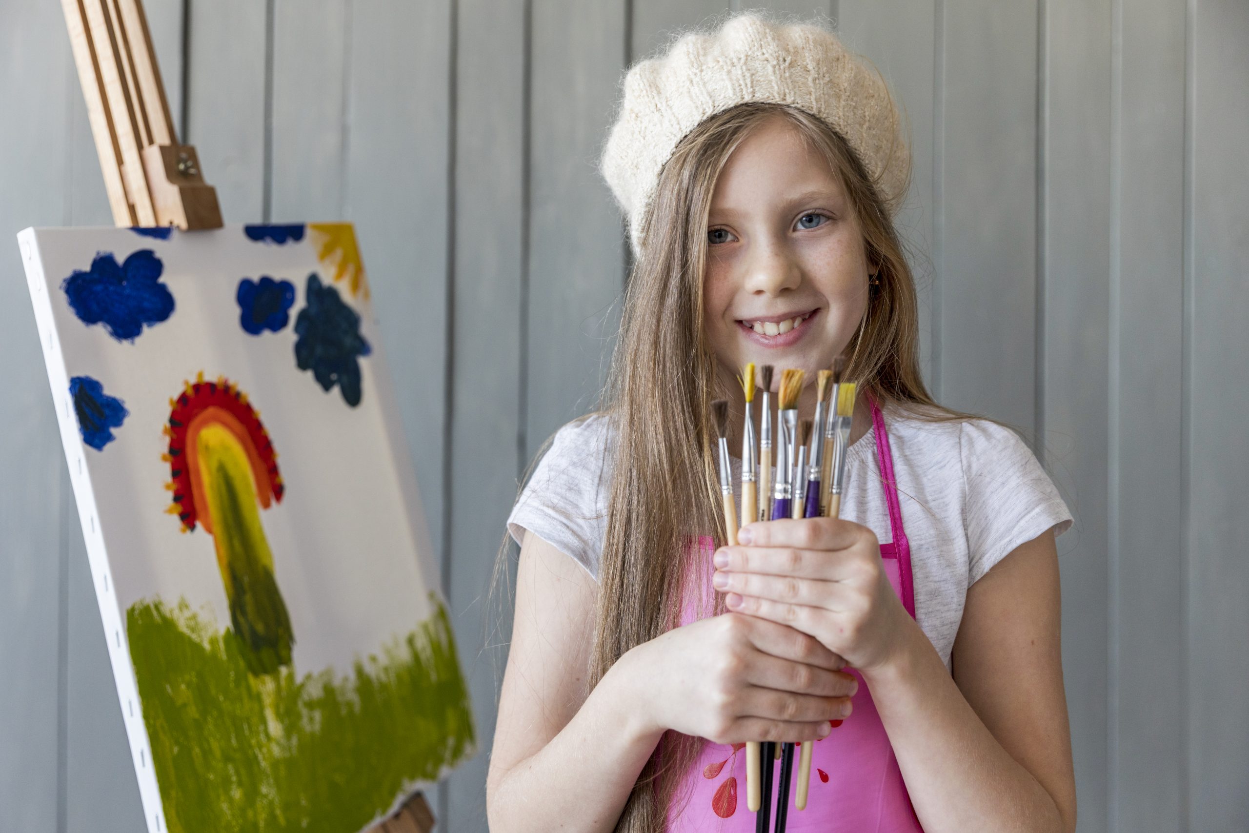 portrait beautiful smiling girl wearing knit cap holding various type brushes standing near canvas
