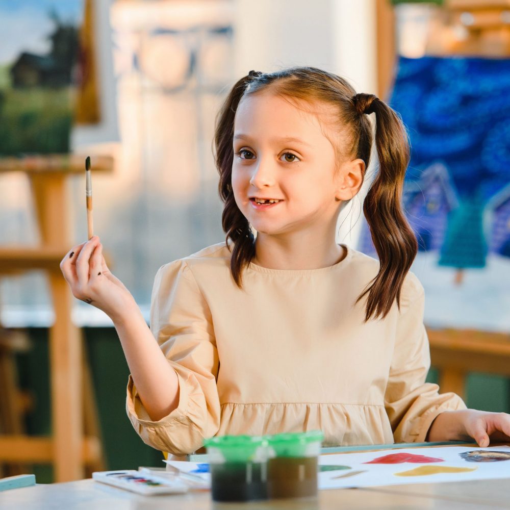 A young girl with pigtails happily paints in an art class with colorful artworks behind her.